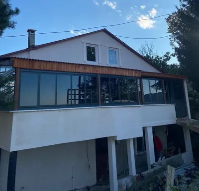 Müstakil evin üst kat terası antrasit alüminyum sürme sistemle baştan başa kapatılmış, ahşap saçak altında uzanan cam cephe